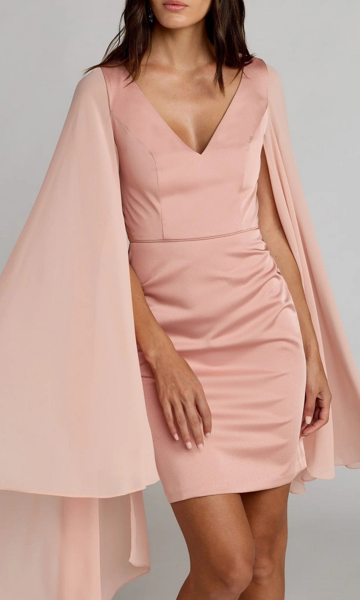 A woman wears the Mac Duggal 55358, a fitted blush pink V-neck short semi-formal dress with elegant chiffon cape sleeves—perfect for any semi-formal occasion.